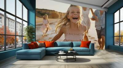 happy family of four running on the beach, a girl in front with blonde hair laughing and smiling wearing a light pink dress Wall mural