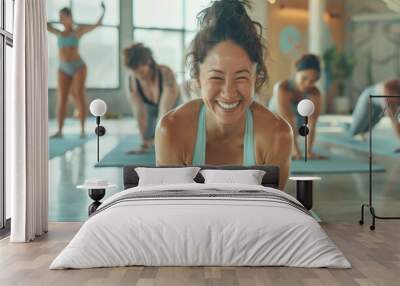 women in yoga attire sitting on black mats laughing during class at the gym, surrounded other students also smiling and doing poses Wall mural