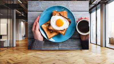 A photo of two hands holding a blue plate with toasts, a fried egg and a coffee cup on a gray wooden table in a top view. Wall mural