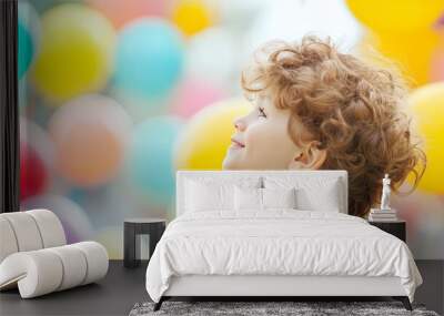 A young boy with curly hair smiles and looks up at colorful balloons in the background. Wall mural