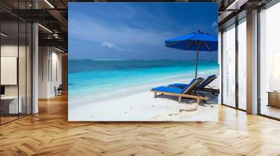 Two lounge chairs under a blue beach umbrella on a white sandy beach with clear blue water. Wall mural