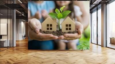 Two miniature houses with a plant growing in the middle are held in the cupped hands of a person. Wall mural