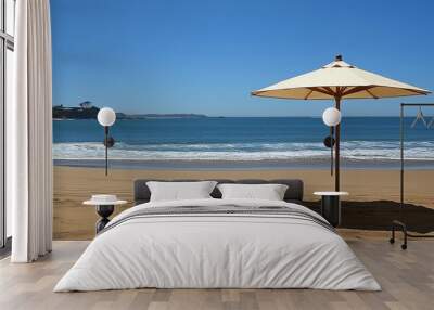 A single white beach umbrella casts a large shadow on the sandy beach. The ocean is calm, blue, and there is a light blue sky. Wall mural