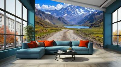 A winding dirt road leads through a mountain valley, with a snow-capped peak in the distance and a sparkling blue lake reflecting the sky. Wall mural