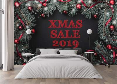 Black stone background with a frame of fir branches decorated with balls and red ribbon. Top view. With text Xmas sale 2019 Wall mural