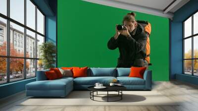 A male traveler takes pictures with a camera while hiking. A tourist with a backpack on his back stands in a studio on a green screen. Concept of travel, active rest, hiking. Wall mural