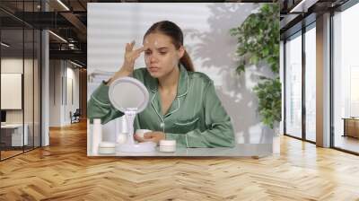 A young woman without makeup applying white cream on problem skin areas of the face. A woman in green silk pajamas sitting at a dressing table in front of a mirror in the morning. Wall mural