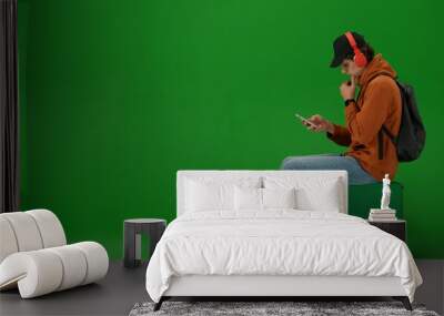 Portrait of person tourist isolated on chroma key green screen background. Young man sitting holding smartphone listening music in headphones. Wall mural