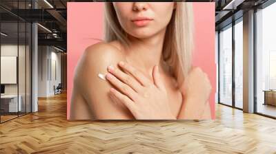 Young blonde woman posing at camera, applying body cream on shoulder skin, isolated on pink studio background close up shot. Beauty skin care concept Wall mural