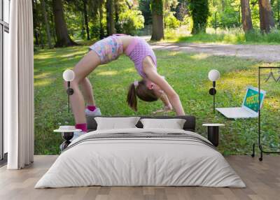 Gymnastic backbend. Teenage girl doing yoga in the park online. Wall mural