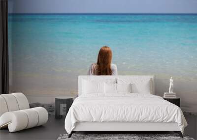 Girl with long red hair sitting with his back on the shore of the blue sea in good weather Wall mural