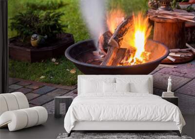 A portable fireplace with bright burning firewoods making sparks and smoke at the backyard  or garden near house. A place for evening meeting and stories Wall mural