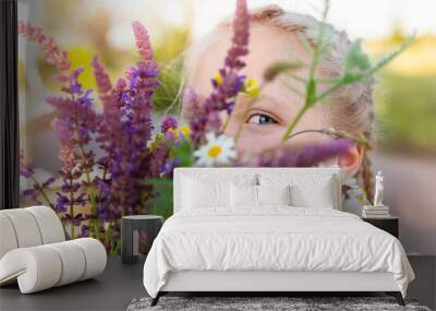 Close-up portrait of little cute caucasian blond kid girl peeking and hiding holding bouquet of salvia sage wild flowers walking at grass meadow outdoors summer day. Young female person wildflowers Wall mural