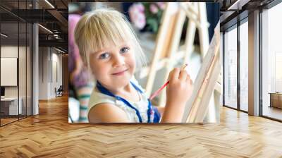 Cute blond smiling girl painting on easel in workshop lesson at art studio. Kid holding brush in hand and having fun drawing with paints. Child development concept Wall mural