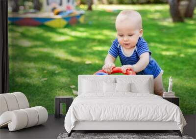 Cute little boy learning to walk with walker toy on green grass lawn at backyard. Baby laughing and having fun making first step at park on bright sunny day outdoors. Happy childhood concept Wall mural