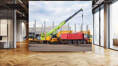 Many different multiclored colorful heavy industrial machinery equipment at construction site parking area against warehouse building city infrastructure development. Commercial vehicles rental sale Wall mural