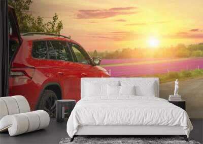 Modern red SUV car parked on rural road near blooming flower field against scenic warm evening sunset light sky landscape. Family countryside vacation auto journey trip at nature outdoors Wall mural