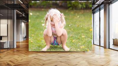 Portrait of cute adorable little blonde caucasian girl squatting on green grass lawn outdoors, having fun and playing hide and seek game, hiding the face with hands. Happy childhood concept Wall mural