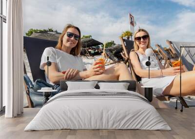 Two young adult happy smiling woman in casual wear enjoy relax sitting in lounger deck chair holding aperol spritz cocktail. Female person drink alcoholic beverage resting lounge area outdoors Wall mural