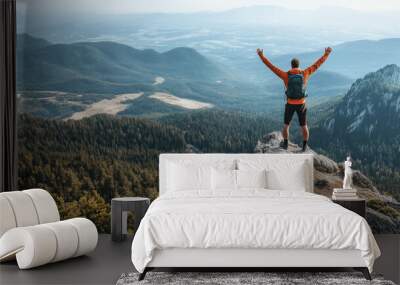 A successful male hiker celebrating on top of a rugged mountain peak, arms raised in victory. The scenic view includes a wide forested valley below and a clear sky. Ai generated Wall mural