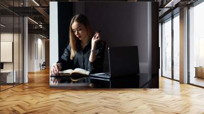 Portrait of serious pretty young business woman wearing stylish black shirt with laptop and diary sitting on workplace and working. Wall mural