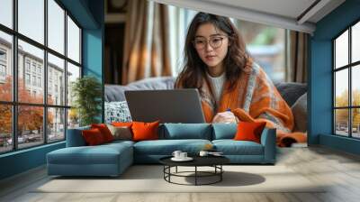 Asian woman wearing glasses writes notes on her notepad while working from home sitting in front of her laptop computer on a sofa in the living room of a house in Edinburgh, Scotland, Generative AI Wall mural