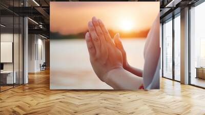 Woman practicing mindfulness meditation in serene sunset by the water with a focus on hands in prayer position Wall mural