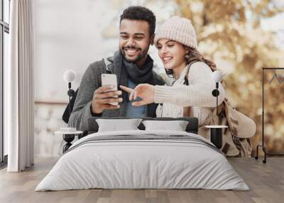 Beautiful happy young couple using smartphone outdoors. Joyful smiling woman and man looking at mobile phone in a city, autumn portrait. Love, technology, communication, winter holiday travel concept Wall mural