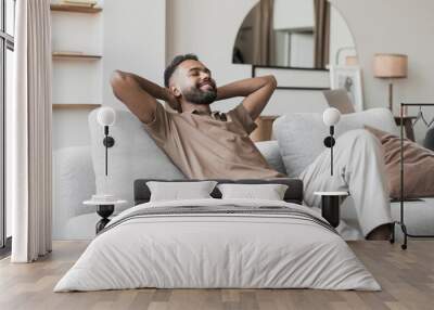 Satisfied handsome young man relaxing on sofa at home in living room, resting after a hard day work, closed eyes, put hands behind head, smiles happily. Relaxation, self care, enjoy life concept Wall mural