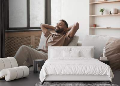 Satisfied handsome young man relaxing on the sofa at home in the living room, resting after a hard day work, closed his eyes, put his hands behind his head, smiles happily Wall mural