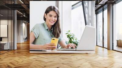 Woman using laptop and smart phone in office. Beautiful girl at desk looking at camera. Entrepreneur, businesswoman, freelance worker, student working on computer. Business, technology concept Wall mural