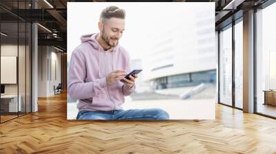 Young handsome men using smartphone in a city. Smiling student man texting on his mobile phone. Coffee break. Modern lifestyle, connection, business concept Wall mural