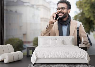 Young handsome student man using smartphone. Smiling joyful guy autumn portrait. Cheerful businessman wearing warm clothes talking by mobile phone in a city Wall mural