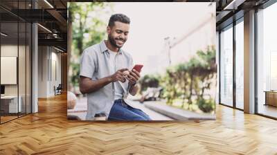 Young handsome student man using smartphone. Smiling joyful guy summer portrait. Handsome man looking at mobile phone Wall mural