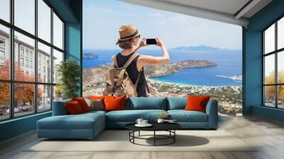 Young tourist woman using smartphone taking photo of beautiful landscape with sea in Greece. Traveler girl with backpack and mobile phone. Travel, tourism, summer holidays, active lifestyle concept Wall mural