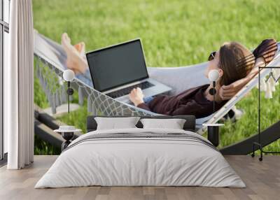 Young woman using laptop outdoor. Young beautiful girl lying in a hammock and working on computer. Freelance, summer, enjoy life, student lifestyle, distance studying and online learning concept Wall mural