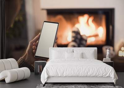 Young woman using smart phone near fireplace at home, white blank empty screen display. Winter lifestyle, leisure, lockdown, holidays, vacations, Christmas time, New Year, communication concept Wall mural