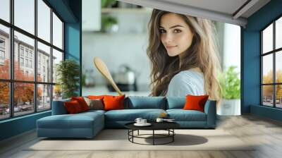 A beautiful woman in a modern kitchen, holding a wooden spoon and looking at the camera with confidence as she gets ready to cook a delicious homemade dish Wall mural