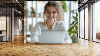 A high-resolution photograph of a young woman marketer, smiling as she works on her laptop in a bright, elegant office setting Wall mural