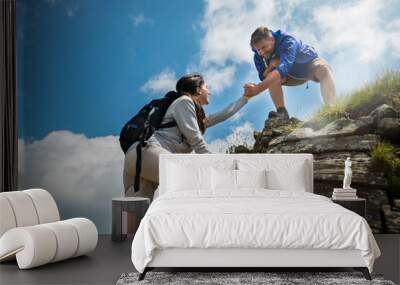 A carefree happy tourists couple is helping to each other to climb on a peak while hiking in the middle of hills surrounded by green nature. Wall mural