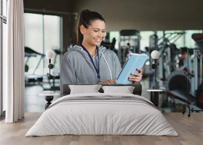 A young personal trainer (girl) holds an Ipad with the gym in the background. Concept: Sport, fitness, happiness. Wall mural