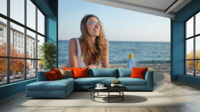 An young smiling woman in white bikini and sunglasses is lying on sandy beach of a seaside with sunscreen or sun tanning lotion anonime bottle behind to take care of her skin during holidays vacation. Wall mural