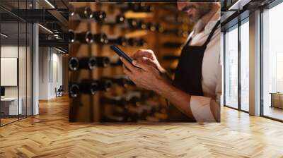 Modern farmer or winemaker is using winery online commerce applications on a smartphone for checking customer service and selling orders summary of his wine production in a wine cellar. Wall mural