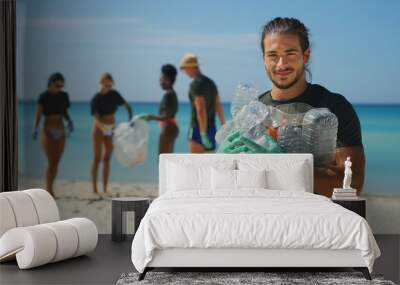 Portrait of an young male volunteer is smiling in camera satisfied with picking up a plastic litter on a beach with a sea to protect an environment. Wall mural