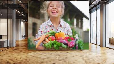 Portrait of happy little girl is holding a basket with fresh biologic just harvested vegetables and smiling in camera on a background of a countryside farm. Wall mural