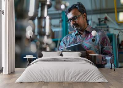 A man in a patterned shirt inspects machinery while holding a tablet in an industrial setting, focusing on engineering equipment operations and maintenance. Wall mural