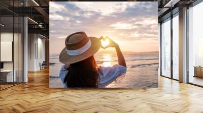 Young woman traveler making heart shape with hands and looking beautiful sunset on the beach, Travel lifestyle concept Wall mural