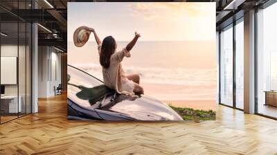 Young woman traveler sitting on a car and looking a beautiful sunset at the beach while travel driving road trip on vacation Wall mural