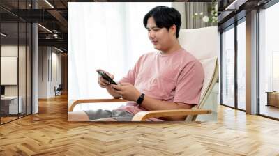 Asian man in casual clothes using smartphone on sofa in living room, working from home or scrolling website. Wall mural