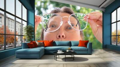 close up female face, funny woman in glasses with metal frame looks through lenses in surprise, concept unexpected object observation, mental human health, regular eye checkups for visual health Wall mural